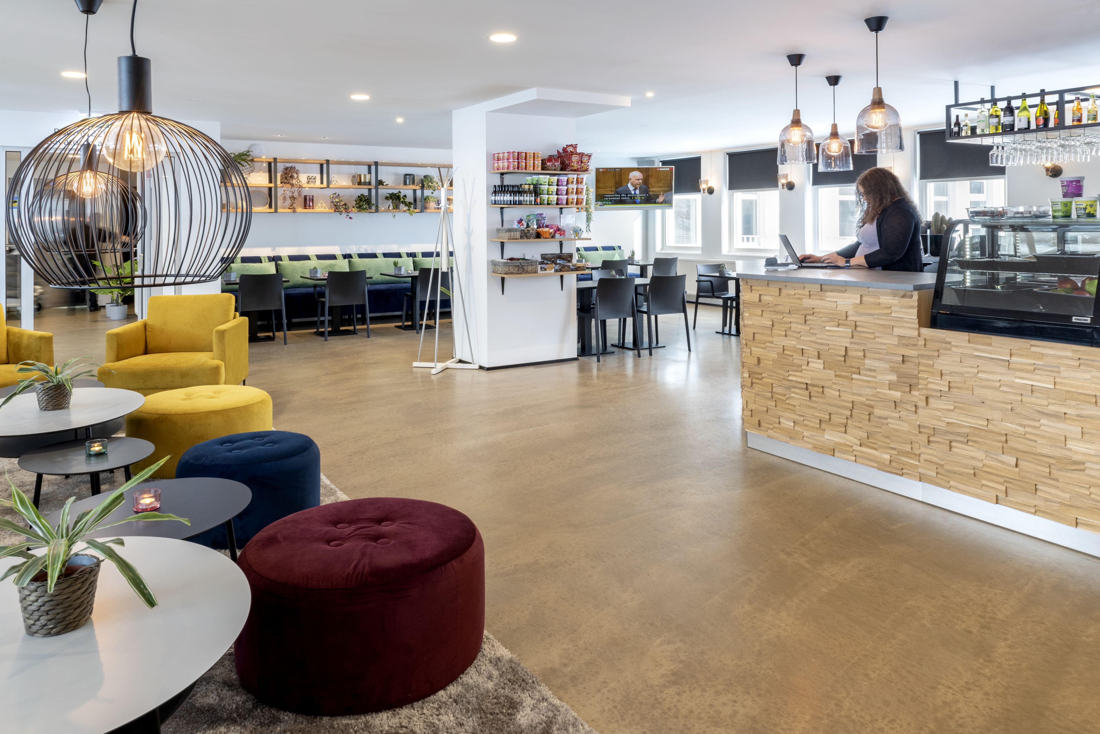 A modern cafe with a wooden counter, seating area with yellow and blue chairs, and a large hanging light fixture.