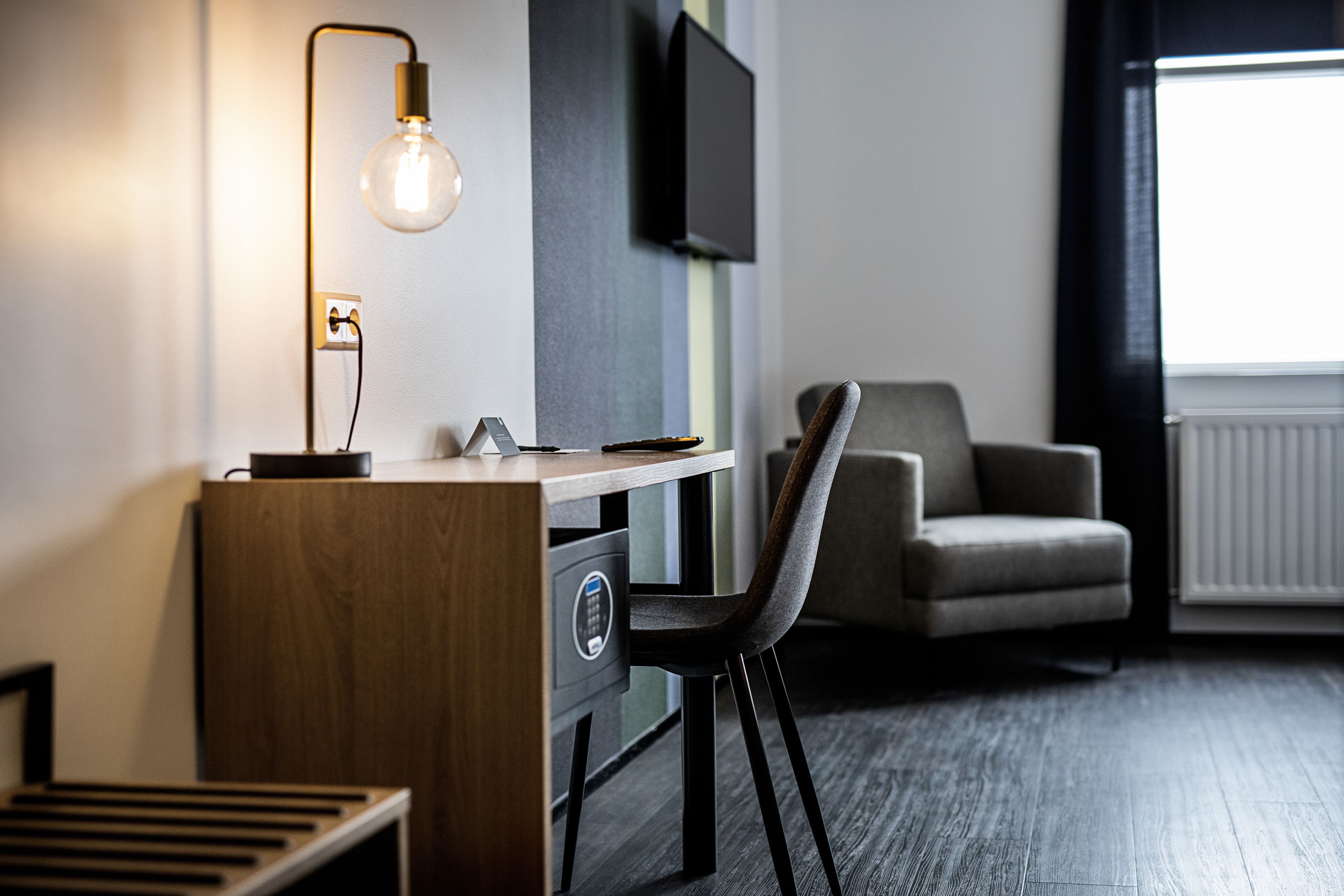 Quarto de hotel moderno com escrivaninha de madeira, cadeira preta, televisão de tela plana na parede e janela com cortinas.