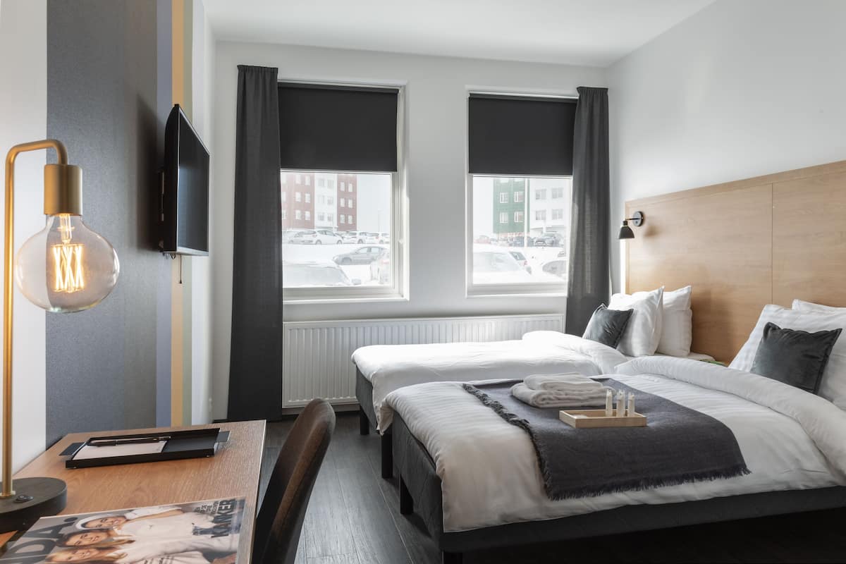 A hotel room with two beds, a desk with a lamp, a TV, and a view of the street through large windows.