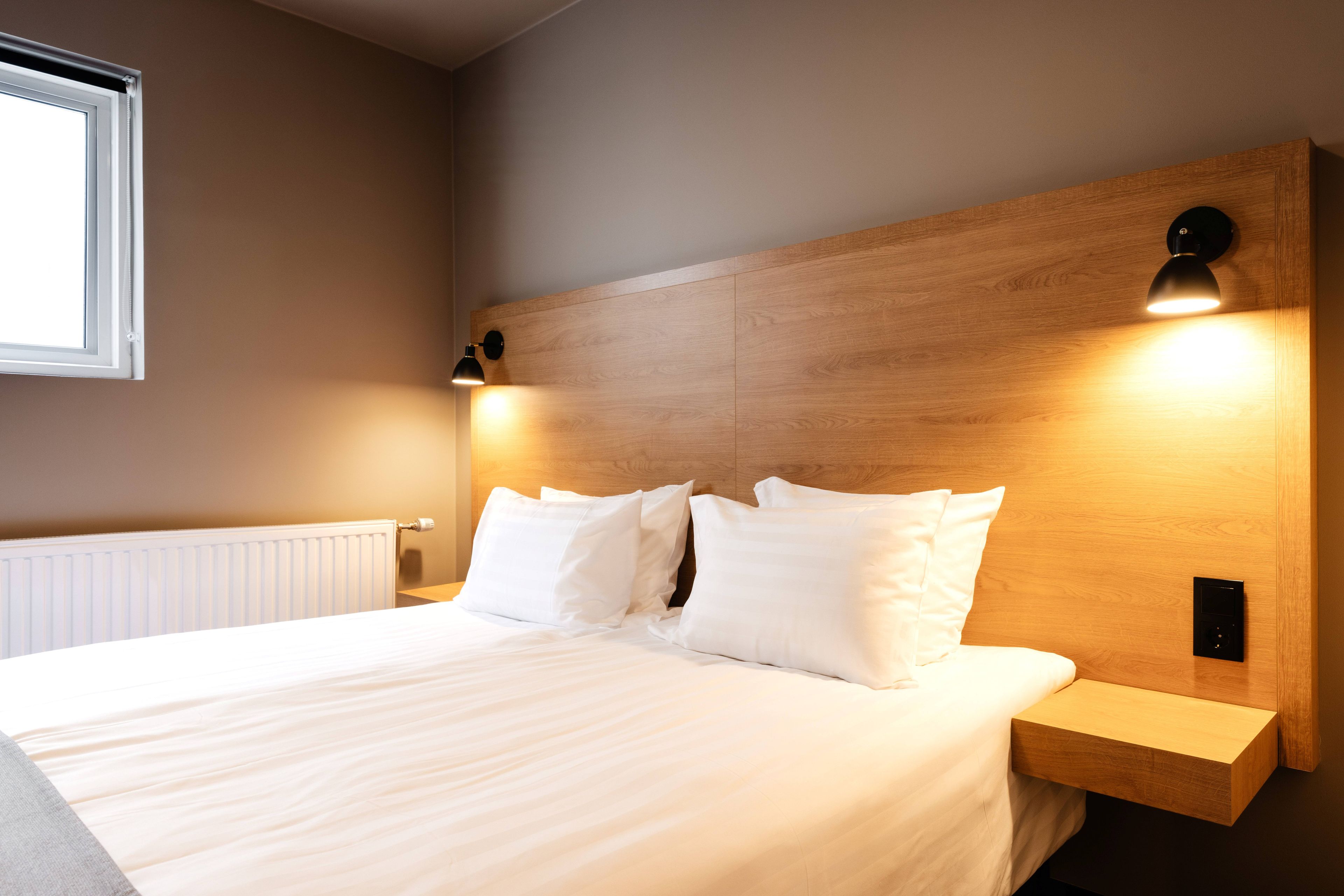 A neatly made bed with white linens and pillows, a wooden headboard, bedside lamps, and a window with blinds.