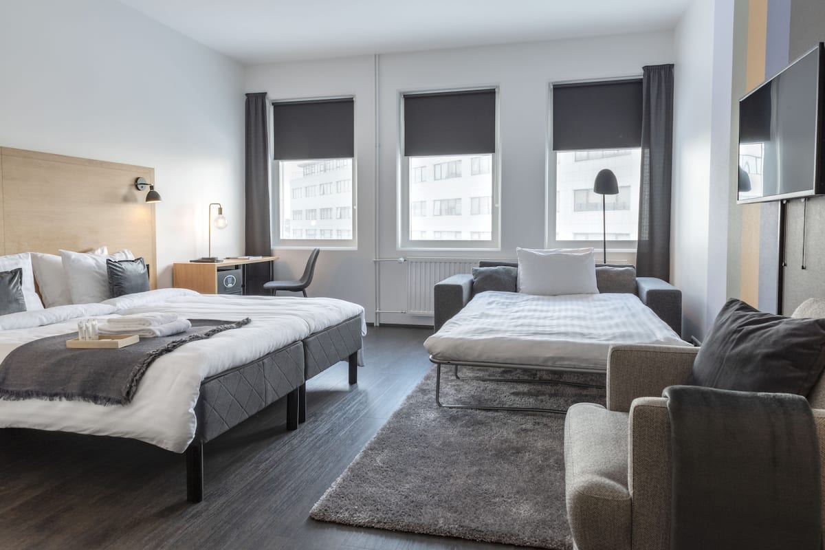 A modern hotel room with two beds, a desk, a chair, a TV, and large windows.