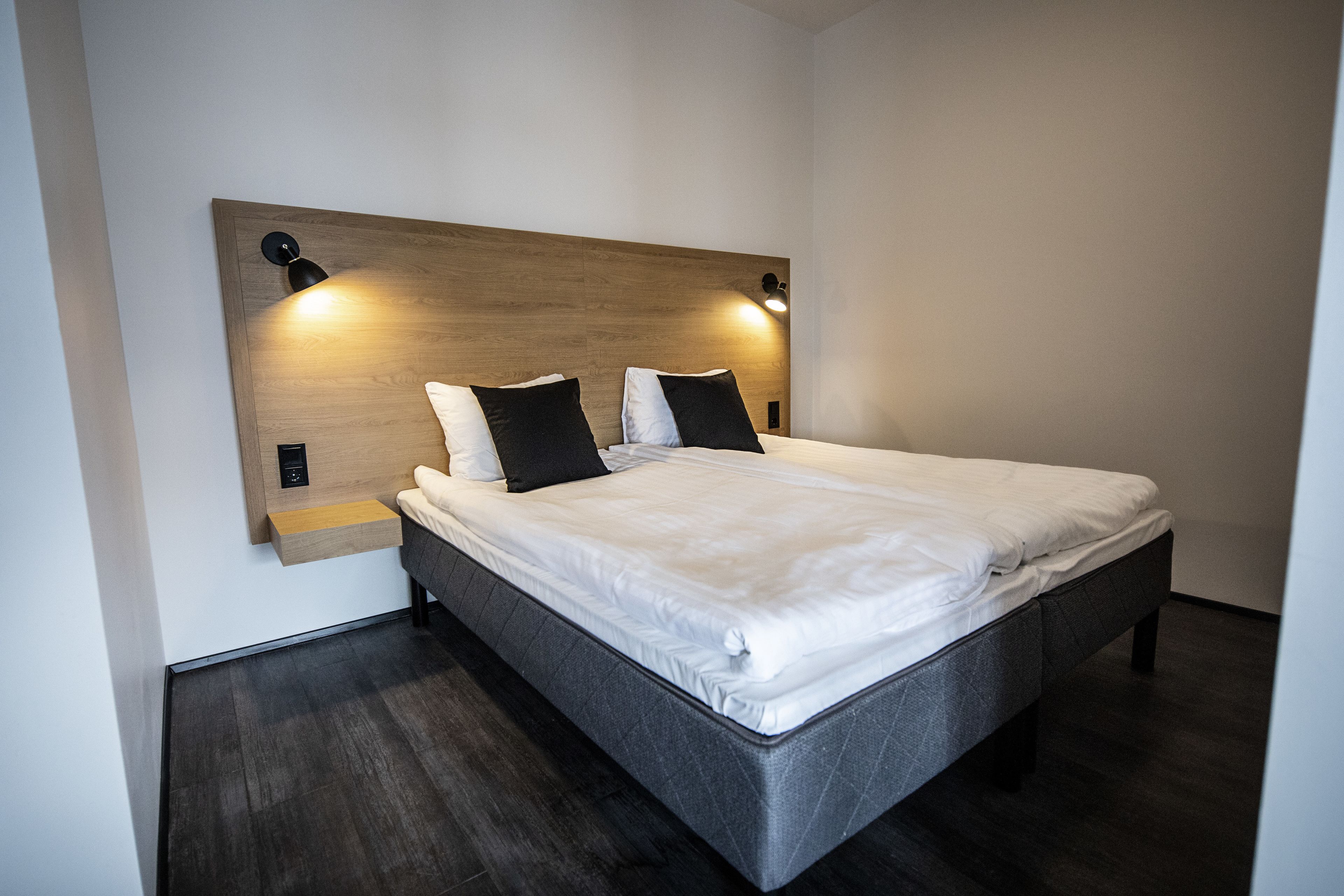 Habitación de hotel moderna con una cama grande, cabecera de madera oscura, dos lámparas de noche y un pequeño estante de madera.