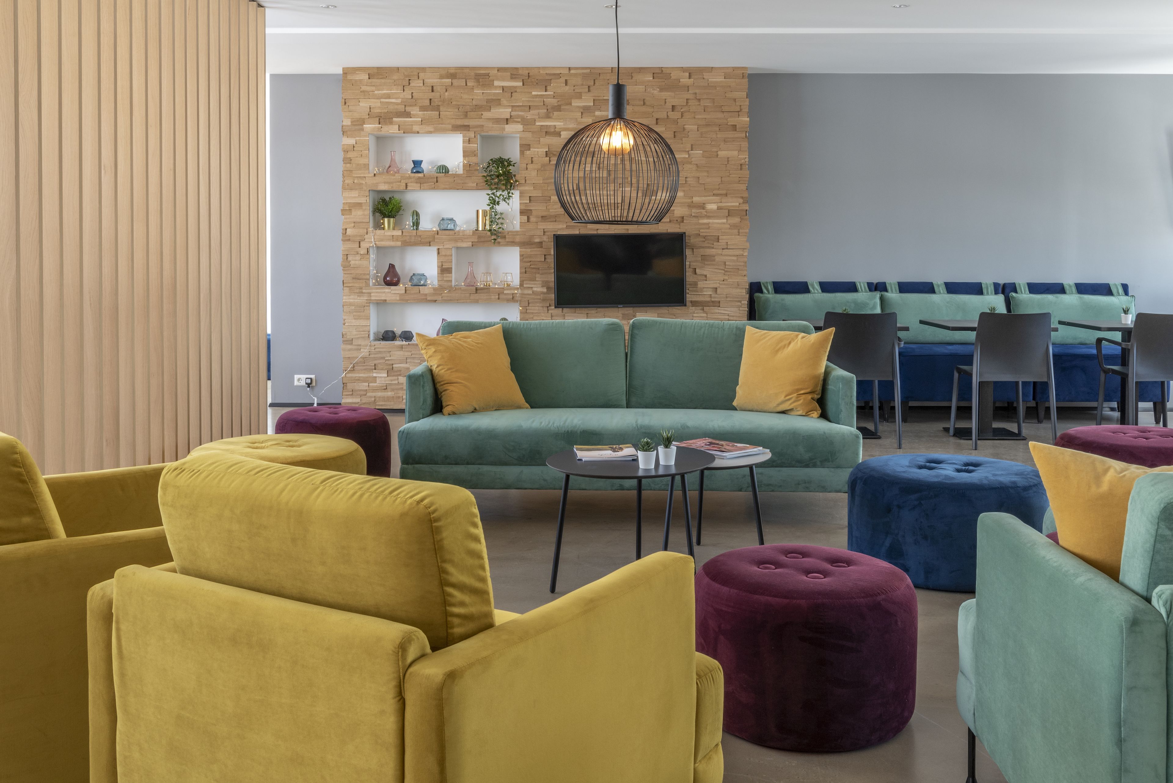 Una sala de estar moderna con un sofá verde, sillones amarillos y una mesa de centro redonda. Se observa una pared de ladrillo con estanterías y un televisor montado en la parte superior.