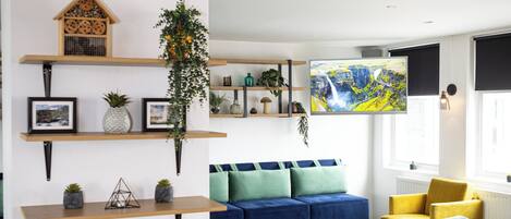 Un salon moderne avec un canapé d’angle, des poufs colorés et des étagères ornées de plantes et d’objets décoratifs.