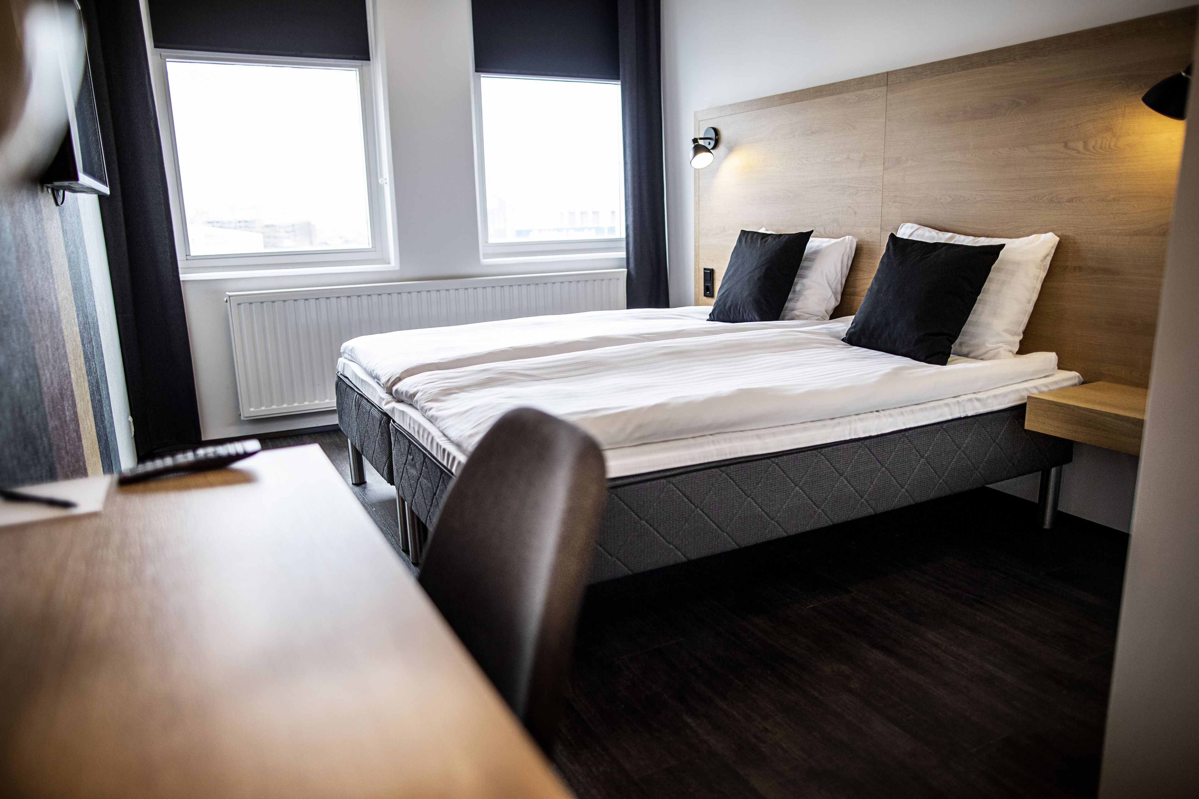 Une chambre d’hôtel moderne avec un grand lit, une tête de lit en bois, une table de chevet et une fenêtre avec des rideaux.