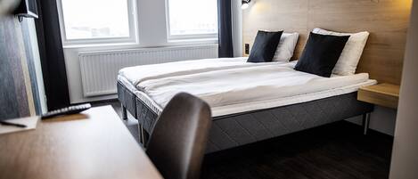A modern hotel room with a large bed, a wooden headboard, a bedside table, and a window with curtains.