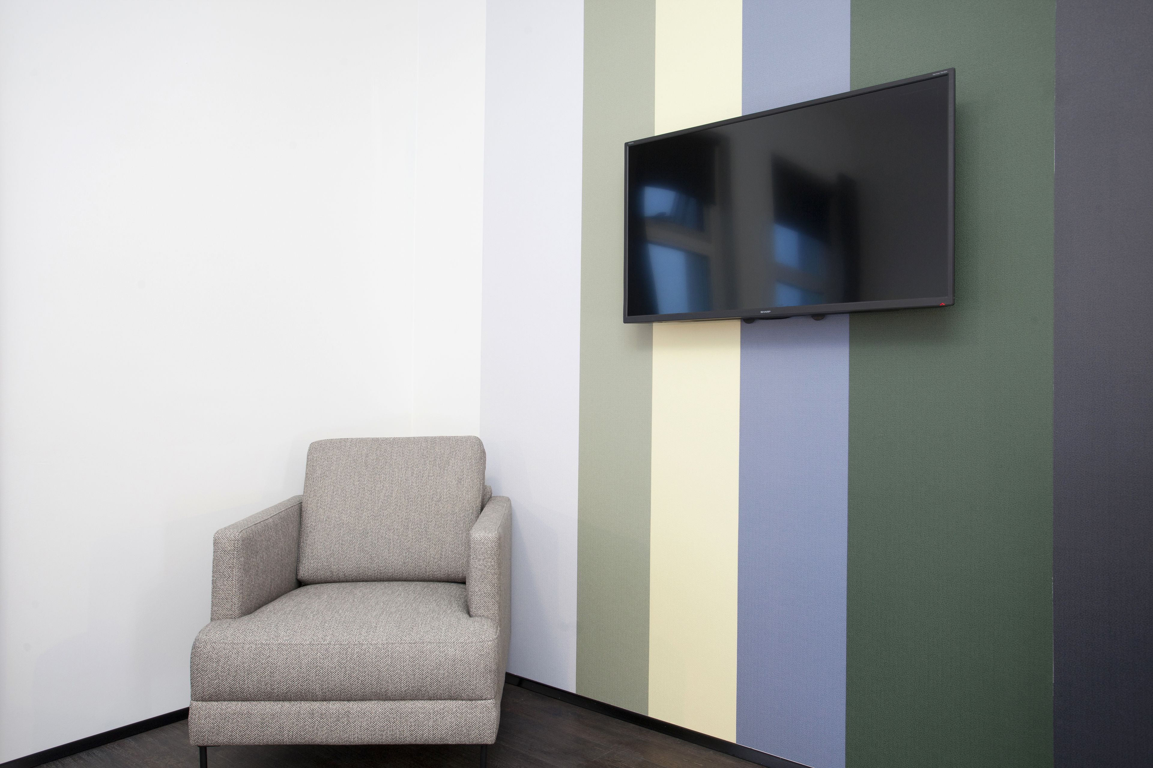 A room with a grey armchair, a flat-screen TV mounted on the wall, and a striped wall with various colors.