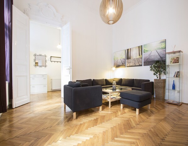 Una sala de estar moderna con piso de madera en espiga, un sofá modular de color oscuro, una mesa de centro de vidrio y una lámpara colgante.