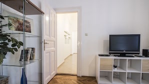 Un salón moderno con un televisor sobre un mueble blanco, una estantería de vidrio con libros y un jarrón azul, y un suelo de madera con patrón de espiga.