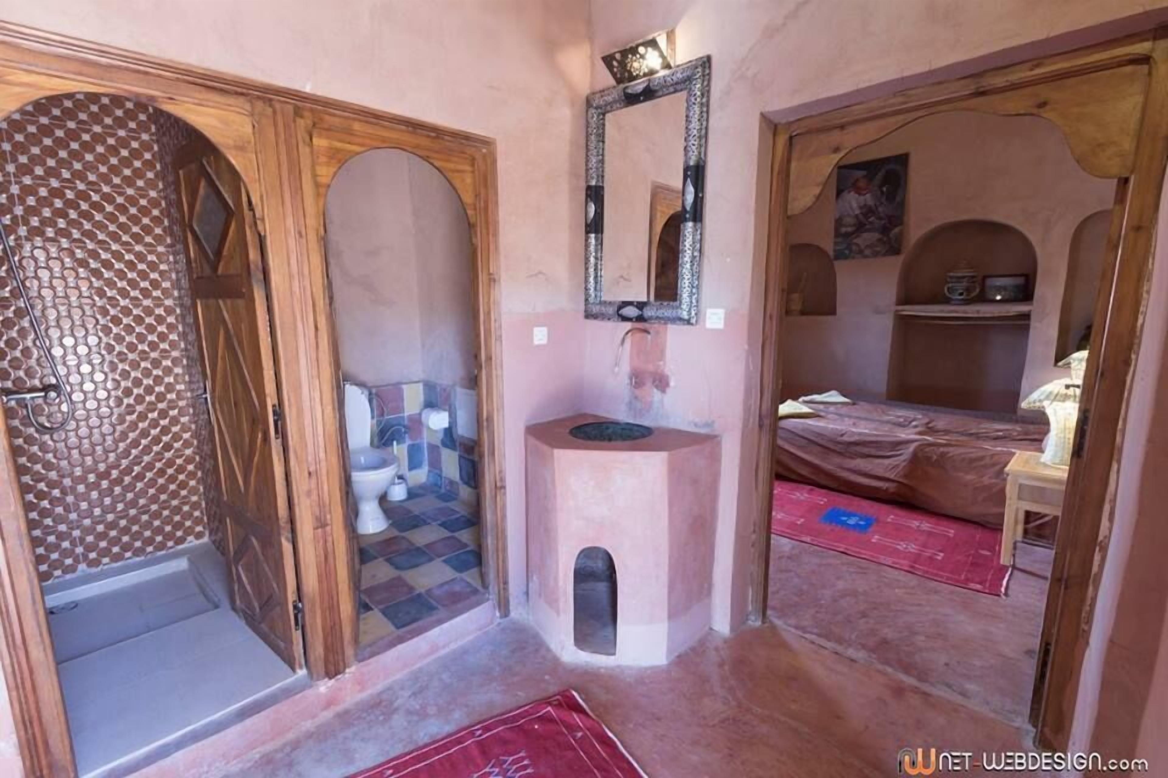 Una habitación con baño, lavabo, espejo, cama y una pequeña estantería.