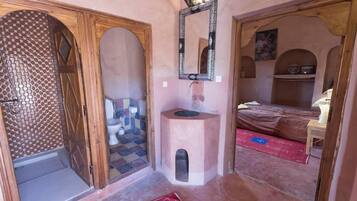 Una habitación con baño, lavabo, espejo, cama y una pequeña estantería.