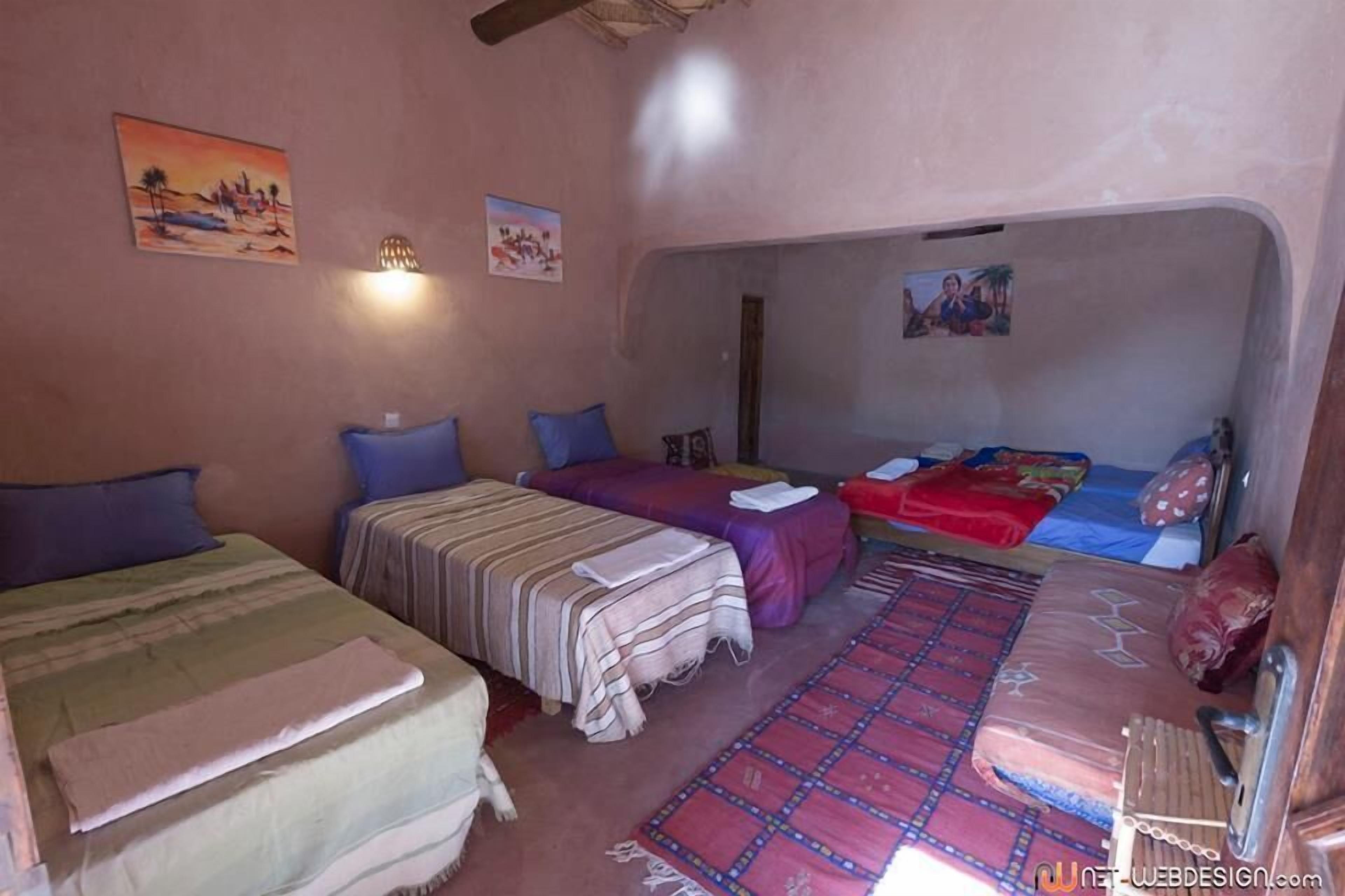 A room with three beds, each with different colored bedding, a patterned rug, and wall art.