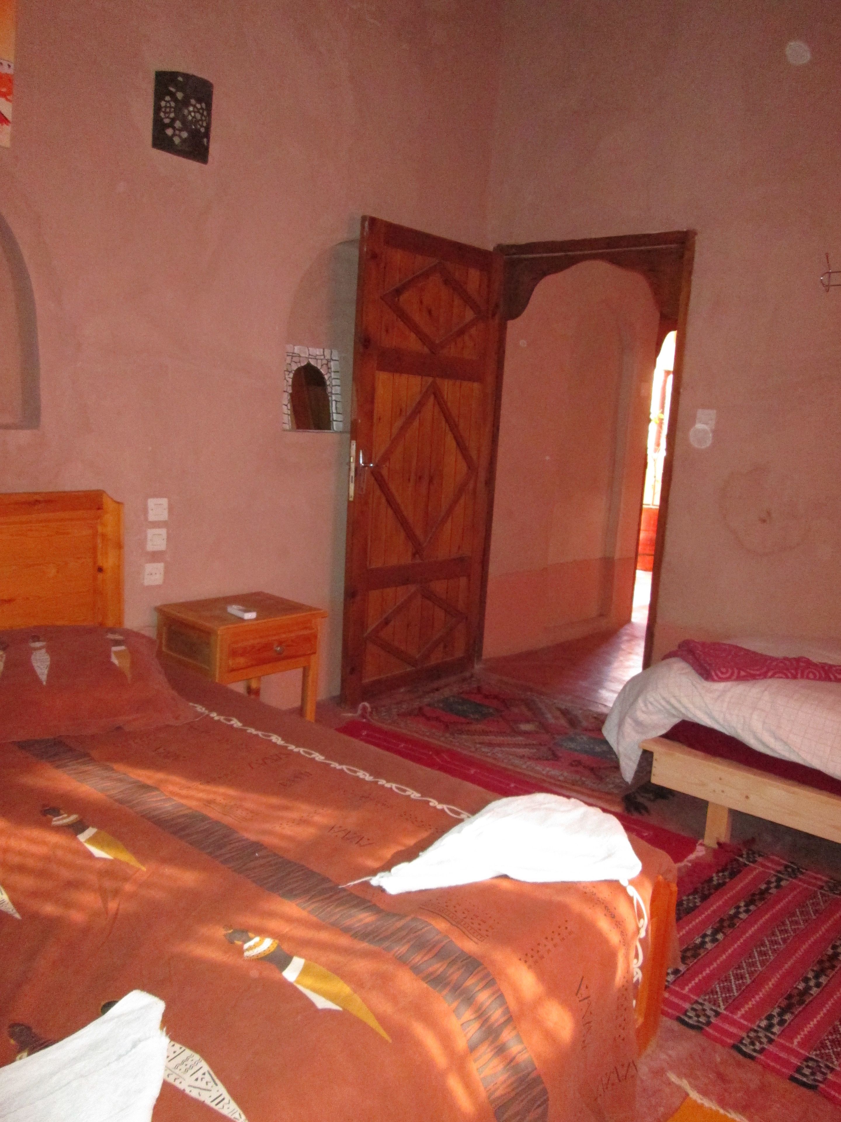 A bedroom with a large bed, a wooden door, a small wooden nightstand, and a patterned rug.