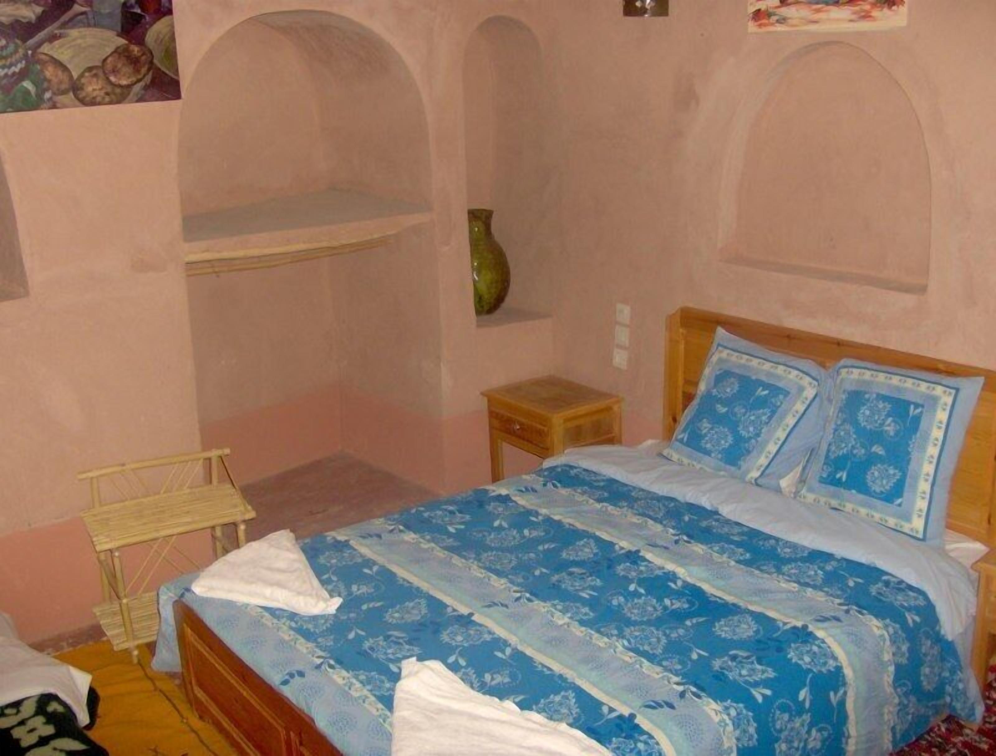 A bedroom with a bed, a small wooden nightstand, a green vase, and a shelf.