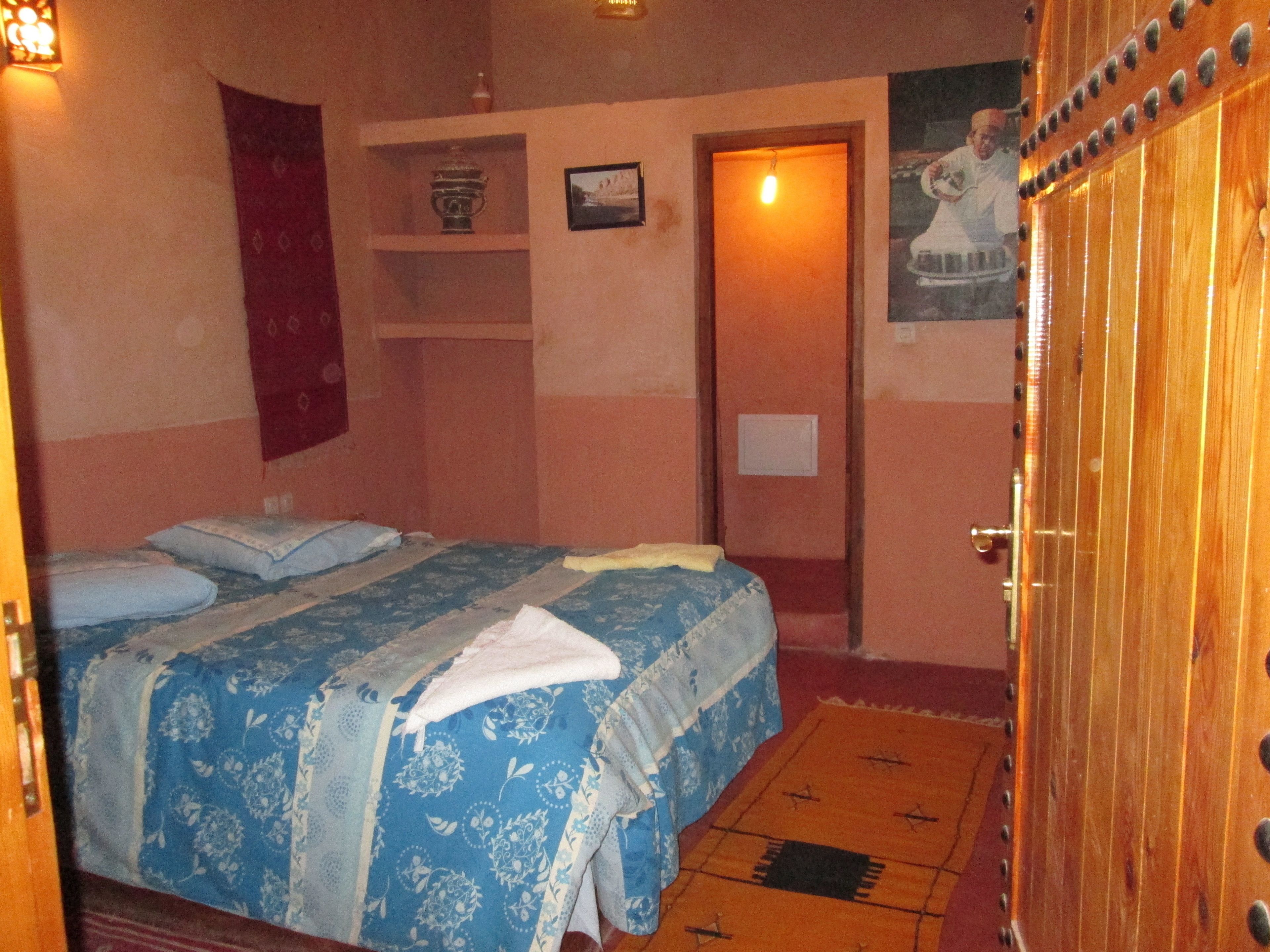 Una habitación con una cama, una puerta de madera, una estantería con un jarrón y un cuadro en la pared.
