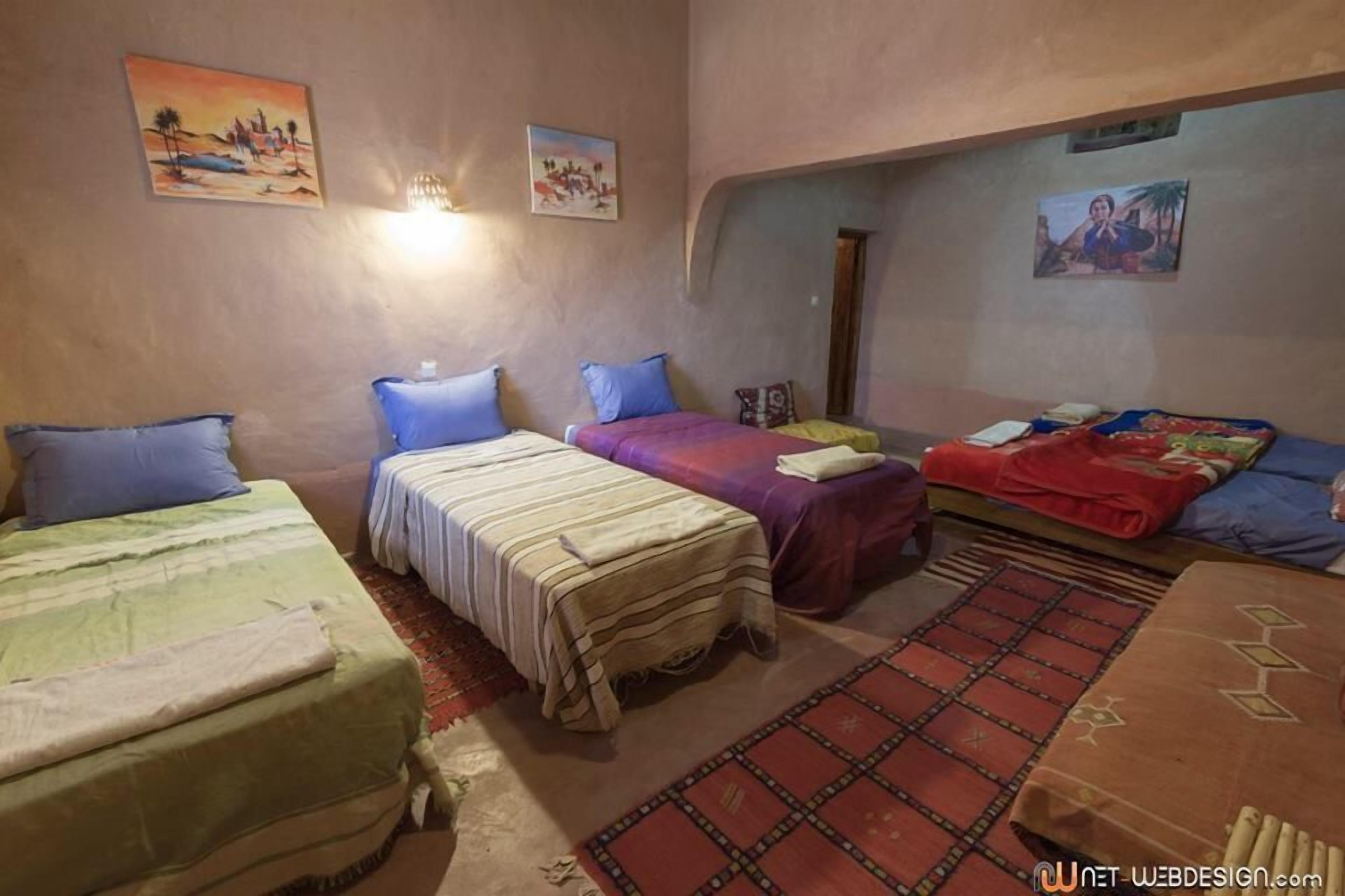 A room with three beds, each with different colored bedding. There are wall-mounted lights and several paintings on the walls.
