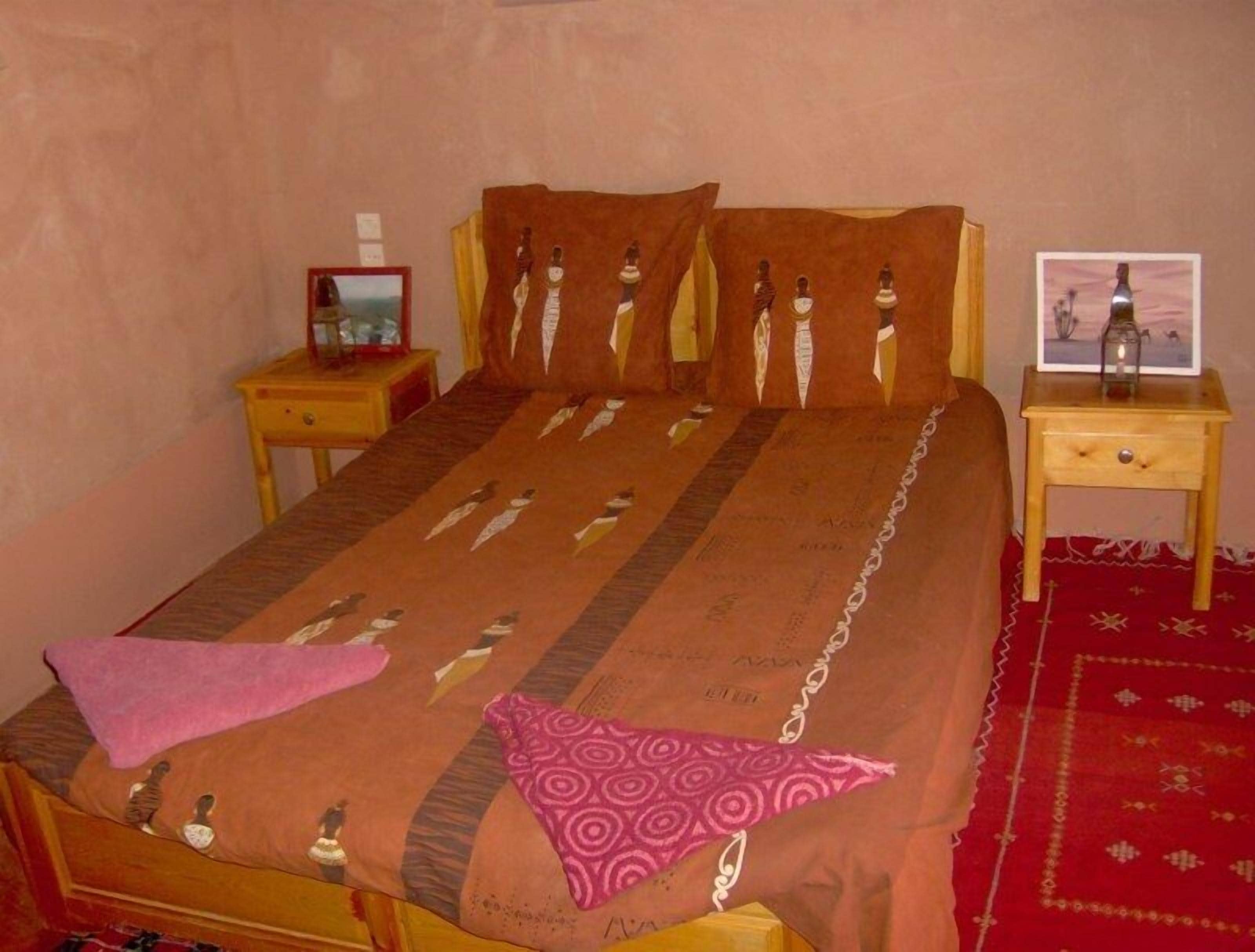 A bed with a patterned bedspread, two pillows, a bedside table with a lamp, and a red rug.