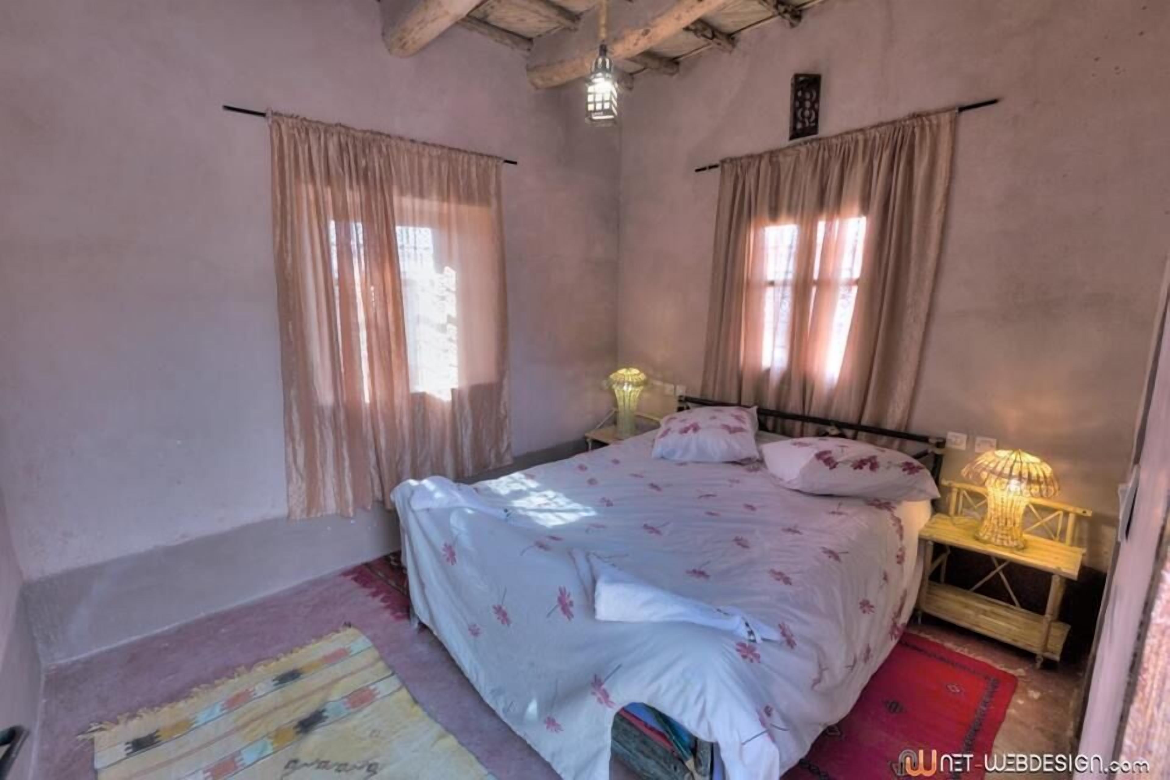 Un dormitorio con cama, cortinas, una mesita de noche y una lámpara.
