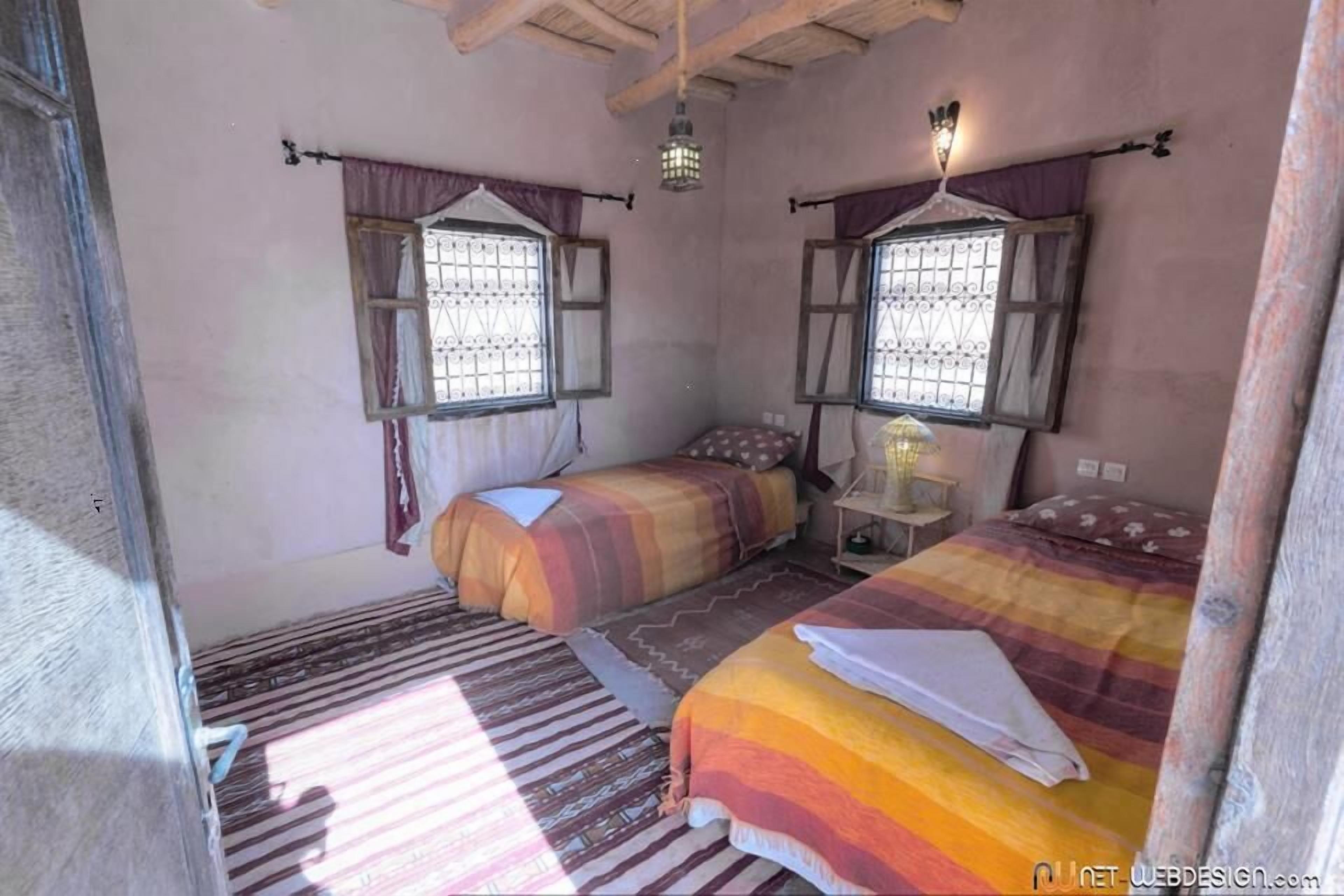 A room with two beds, each with a striped bedspread, a nightstand, a lamp, and a window with a curtain.