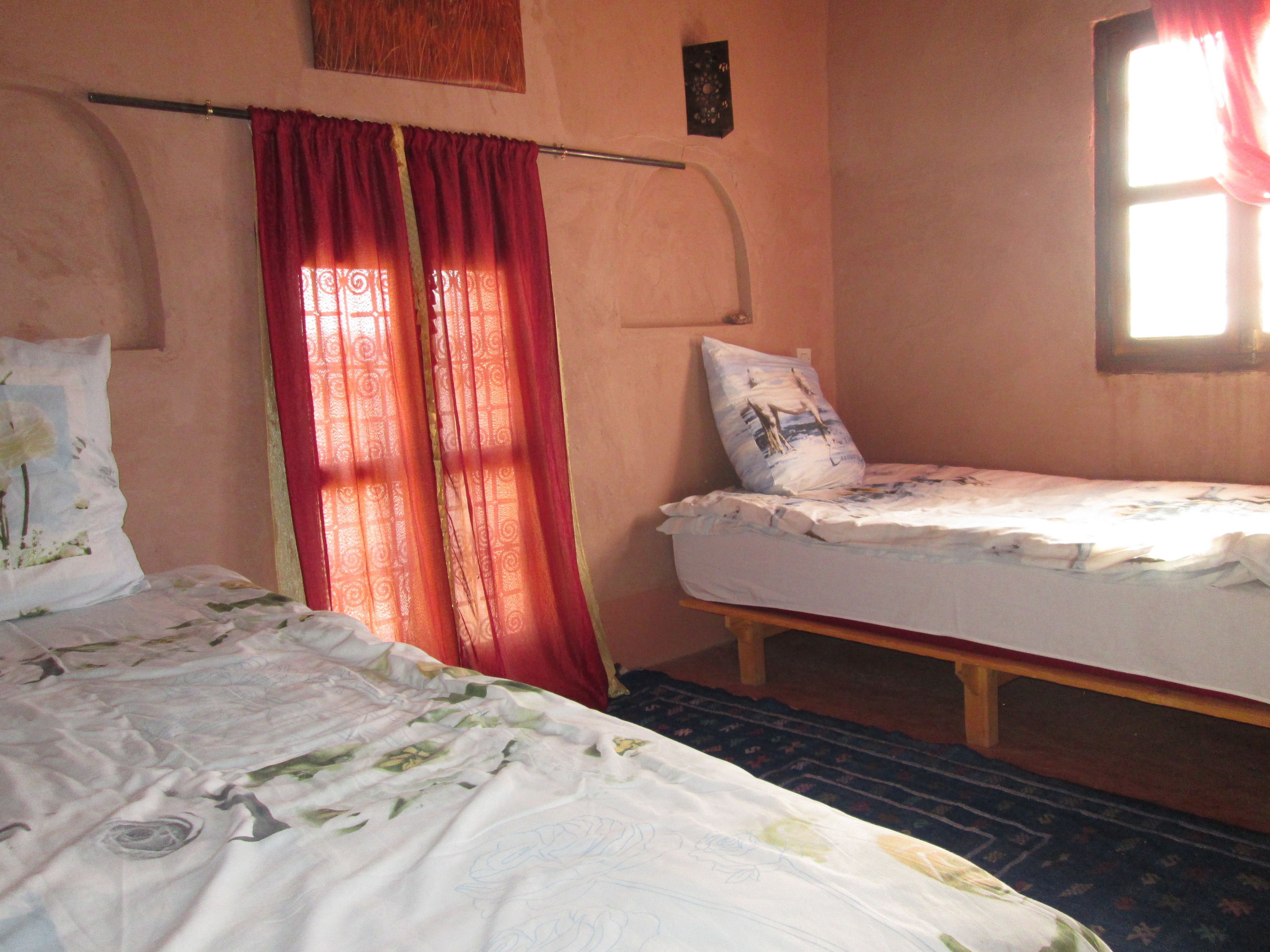 A room with two beds, one with a floral bedspread and the other with a plain sheet. There is a window with red curtains, a wall clock, and a small picture frame.
