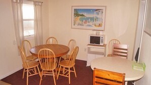 A dining area with a round table and six wooden chairs, a white cabinet with a microwave, and a painting on the wall. - Eagle Bay 204 (Ocean City)