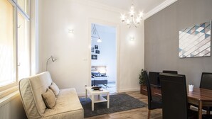 Una sala de estar moderna con un sofá, una mesa de comedor con sillas, una araña y vistas al dormitorio.