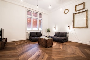 A modern living room with a herringbone wood floor, two dark sofas, a glass-top coffee table, and a large window with a view.