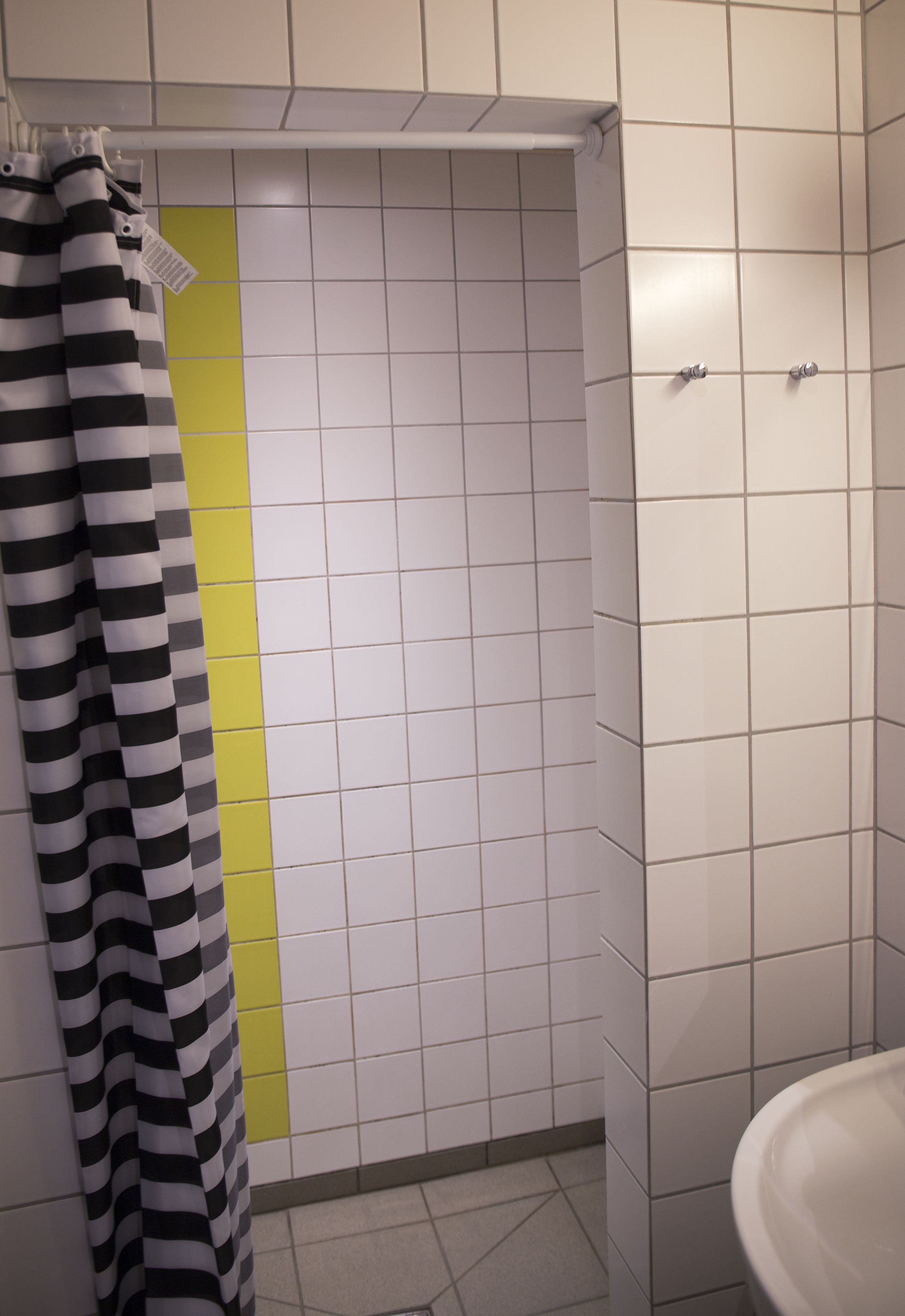 Een badkamer met witte betegelde wanden, een gele verticale streep, een gestreept douchegordijn en een witte wastafel.