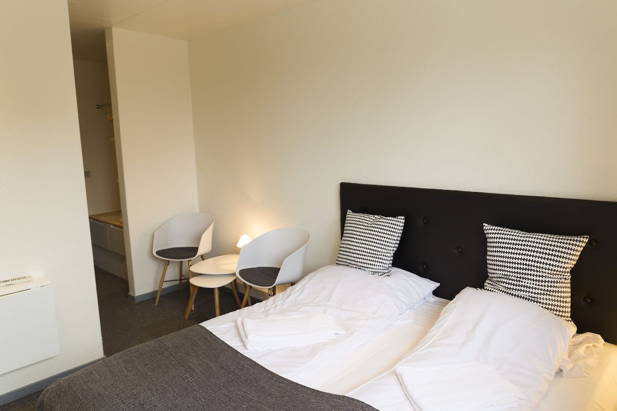 Een hotelkamer met een bed, twee witte stoelen, een zwart hoofdeinde en twee kussens met een patroon.