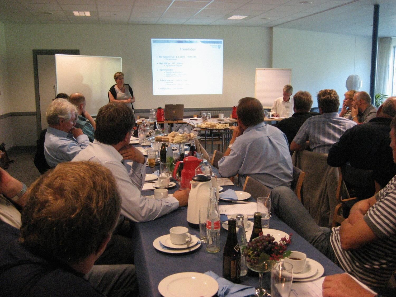 A conference room with a presentation screen, tables with plates, cups, and bottles, and attendees seated around.
