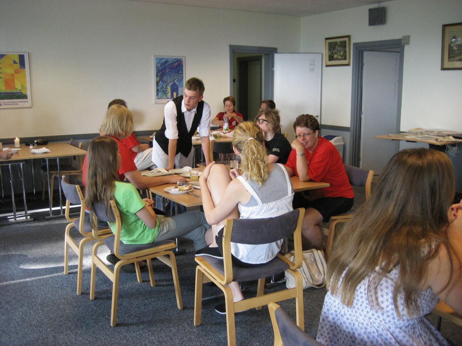 A group of people seated around tables in a room with chairs, a whiteboard, and framed pictures on the wall.