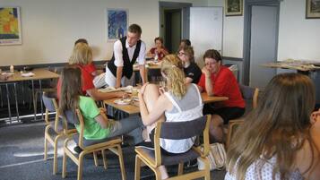 A group of people seated around tables in a room with chairs, a whiteboard, and framed pictures on the wall.