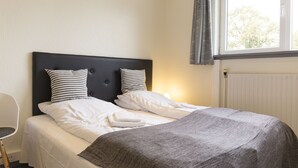Un lit bien fait, recouvert d’une couverture grise, avec deux coussins à motifs et du linge de lit blanc, dans une pièce dotée d’une fenêtre et d’un radiateur.
