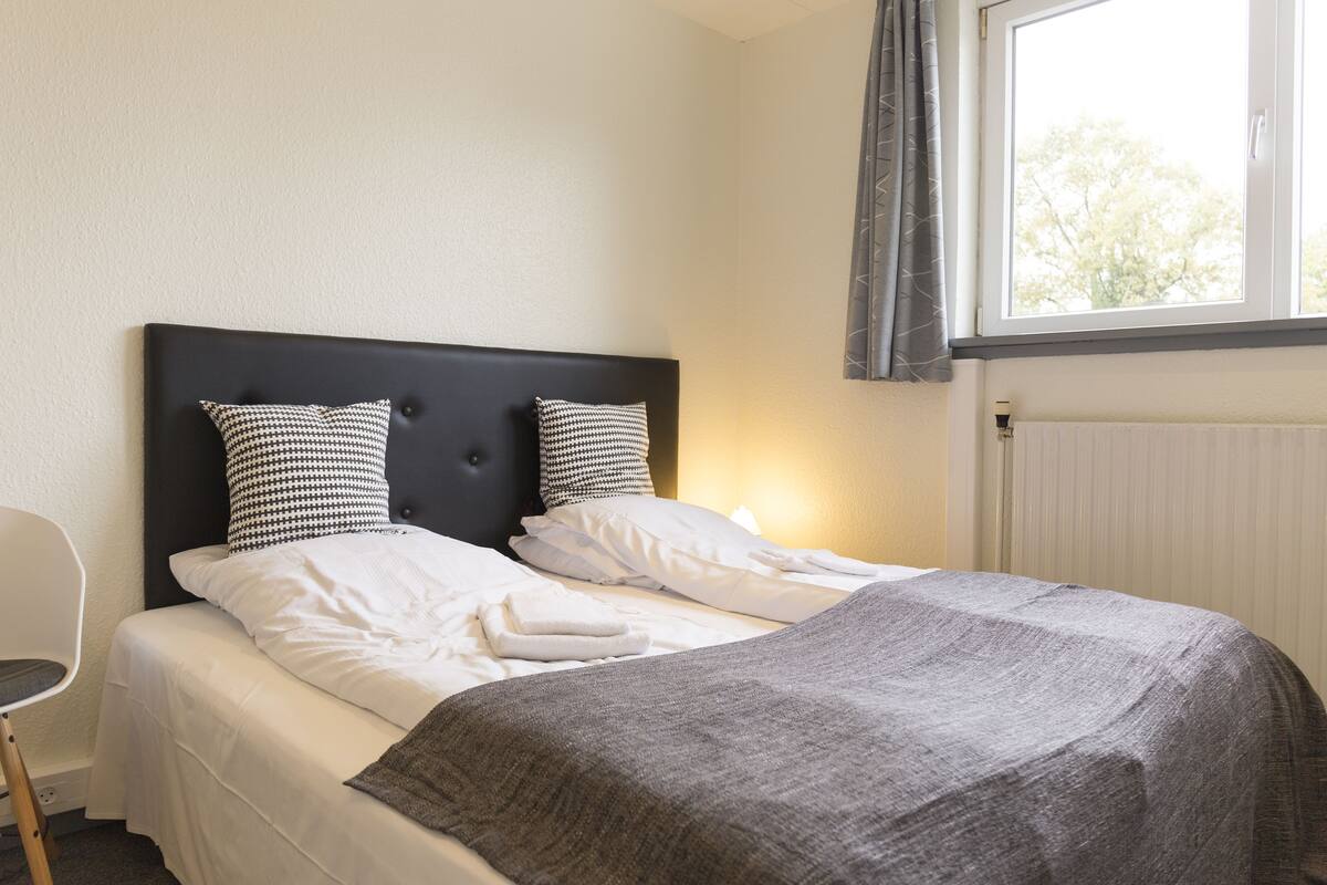 Een net opgemaakt bed met een grijze deken, twee gestreepte kussens en witte lakens in een kamer met een raam en een radiator.