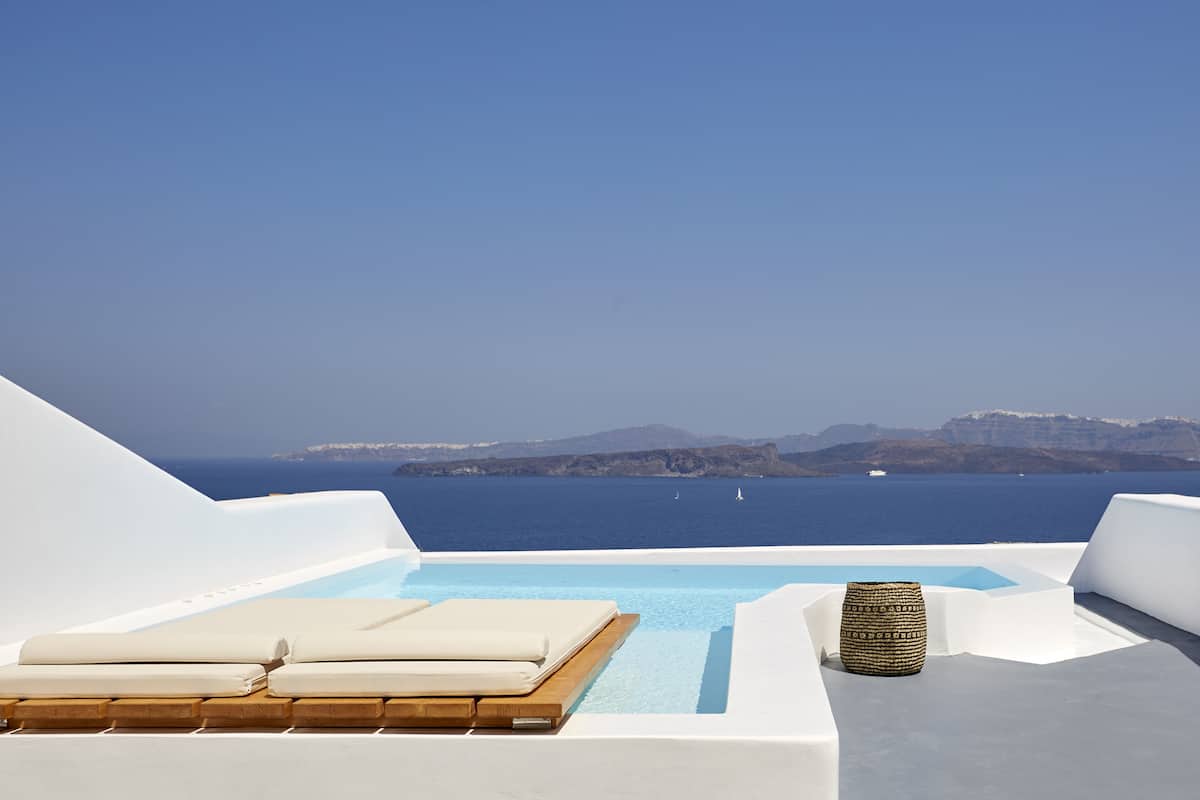 Une piscine à débordement située sur le toit, avec des chaises longues blanches et des coussins à motifs, offrant une vue imprenable sur une mer bleue et des falaises lointaines.