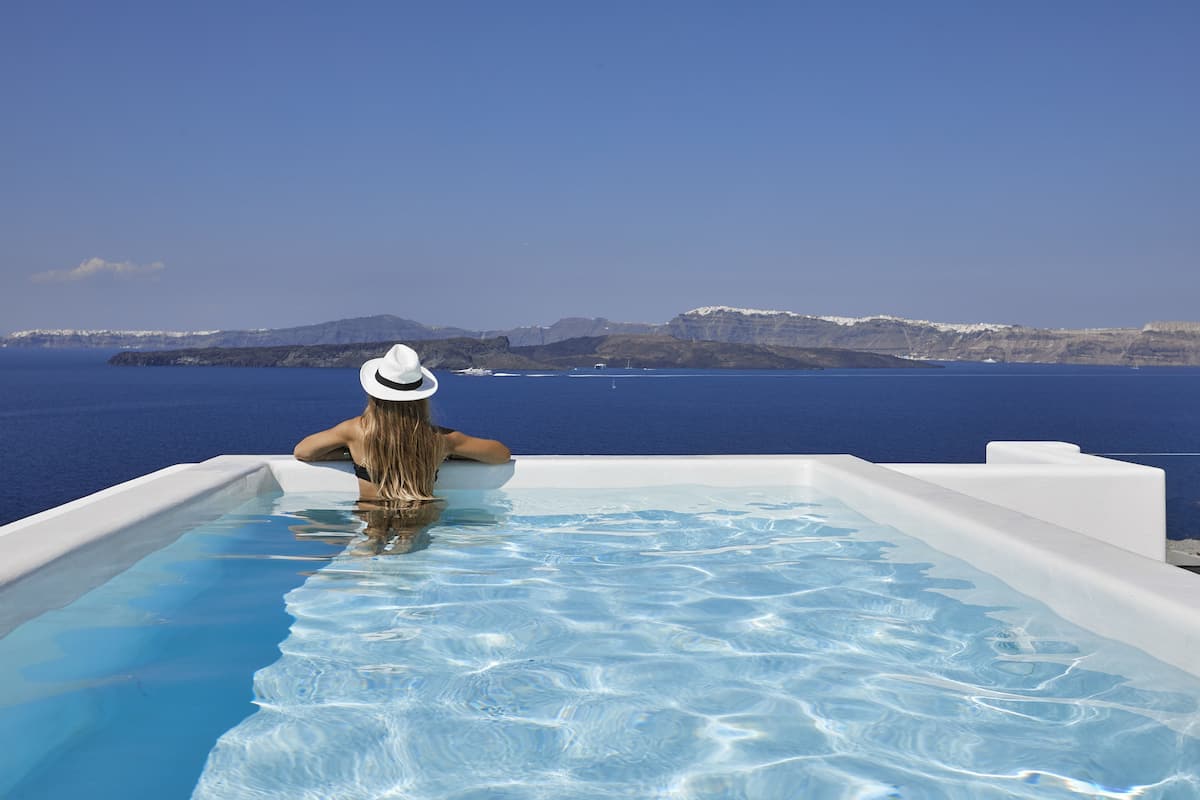 Une personne se détend dans une piscine à débordement aux eaux d’un bleu limpide, avec une vue sur une côte montagneuse.