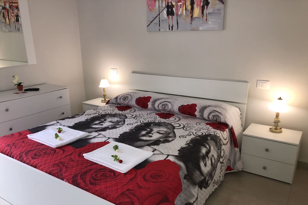 Un lit bien fait, recouvert d’une couvre-lit à motifs floraux rouges et noirs, avec deux lampes de chevet et une petite plante en pot posée sur la table de chevet.