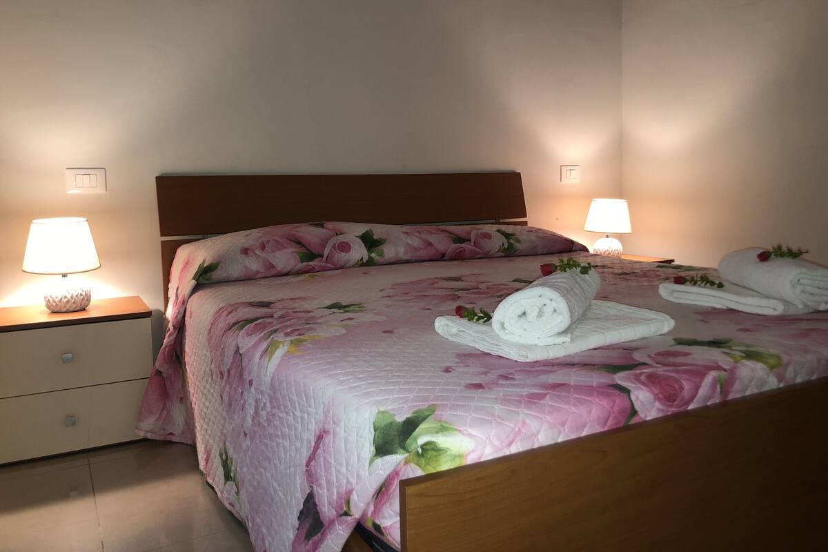 Un lit bien fait, avec une literie à motifs floraux, deux lampes de chevet et des serviettes roulées décorées de roses.