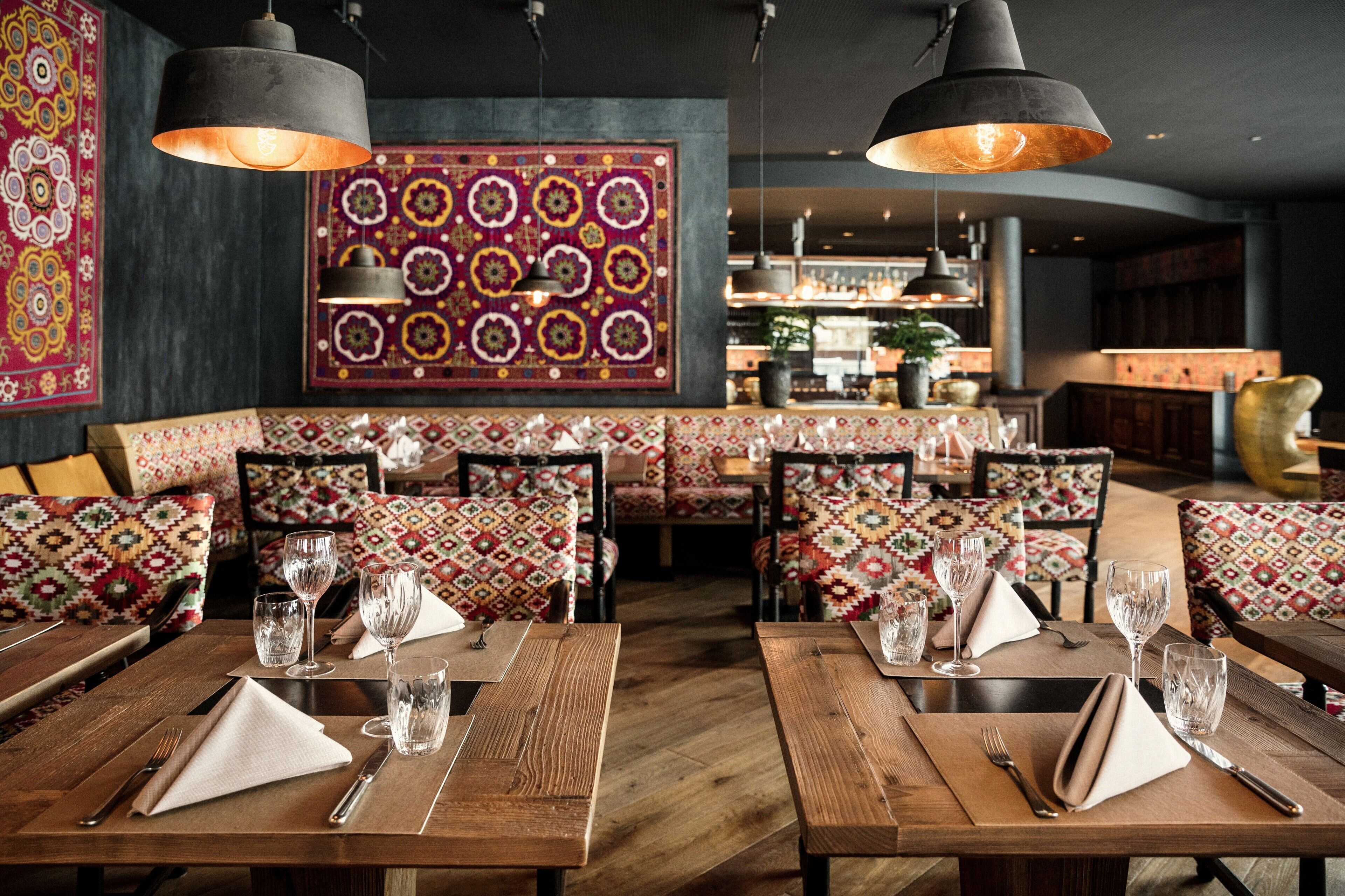 A restaurant with wooden tables and benches, patterned cushions, and hanging pendant lights.