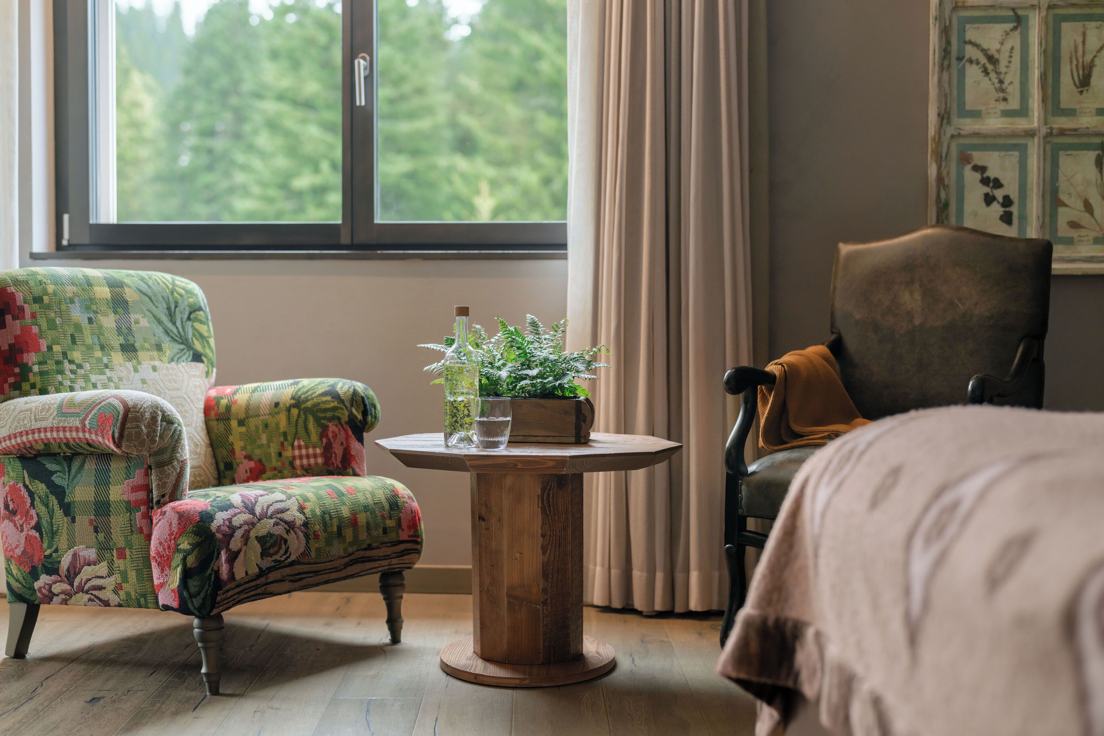 A room with a large window, a round wooden table with a plant, two patterned armchairs, and a plain chair.