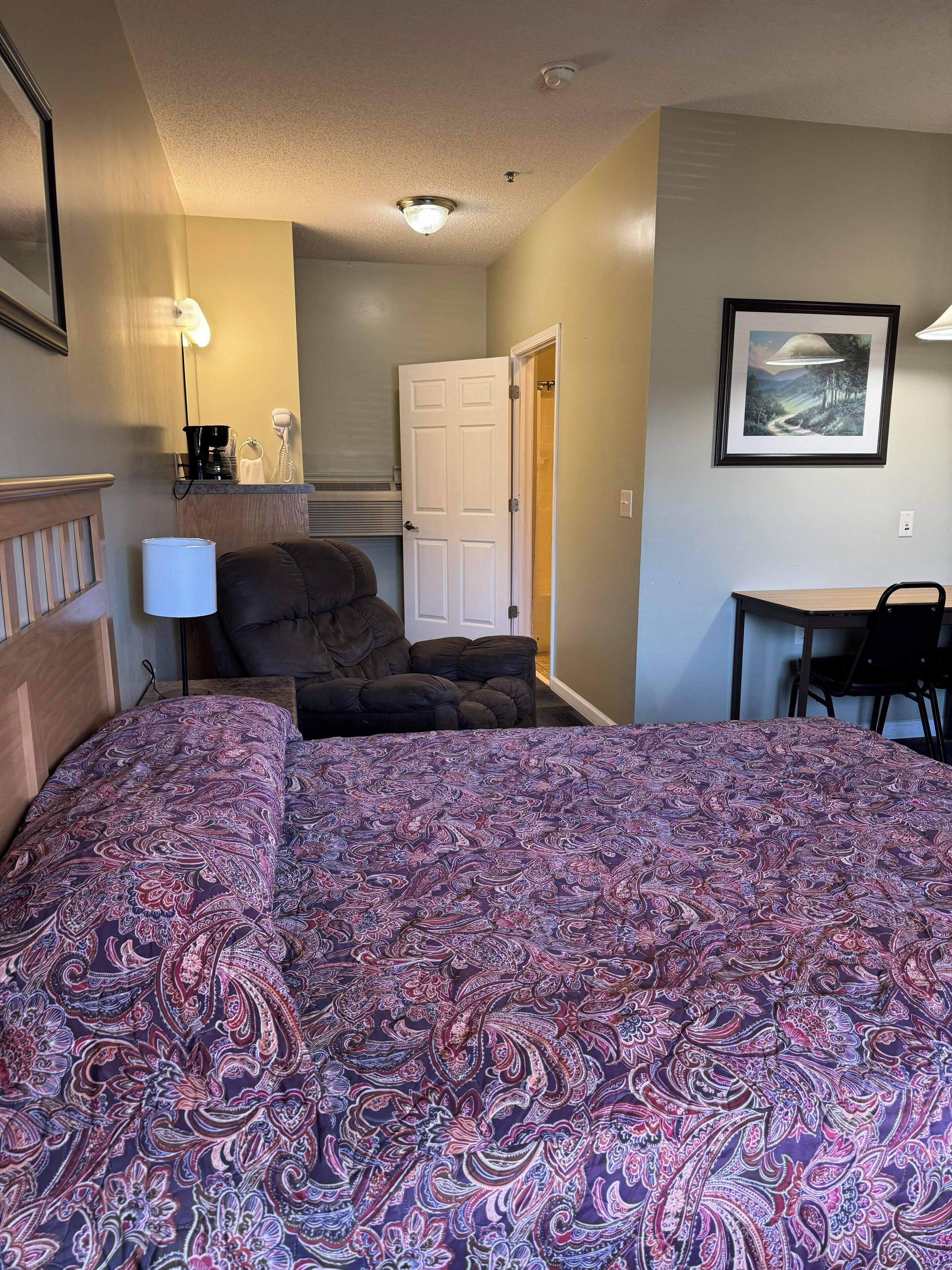 A hotel room with a bed, a chair, a desk, a door, and a framed picture on the wall.