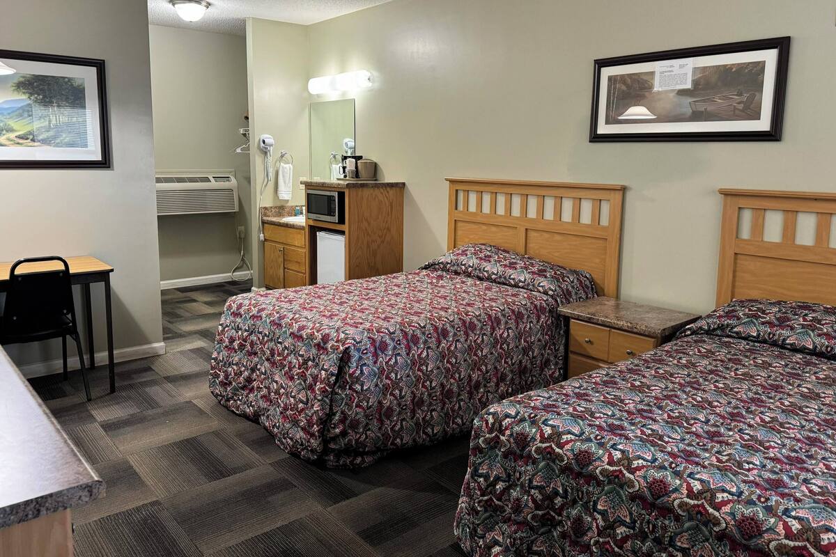 A hotel room with two beds, a desk, a chair, a microwave, a sink, and a mirror.