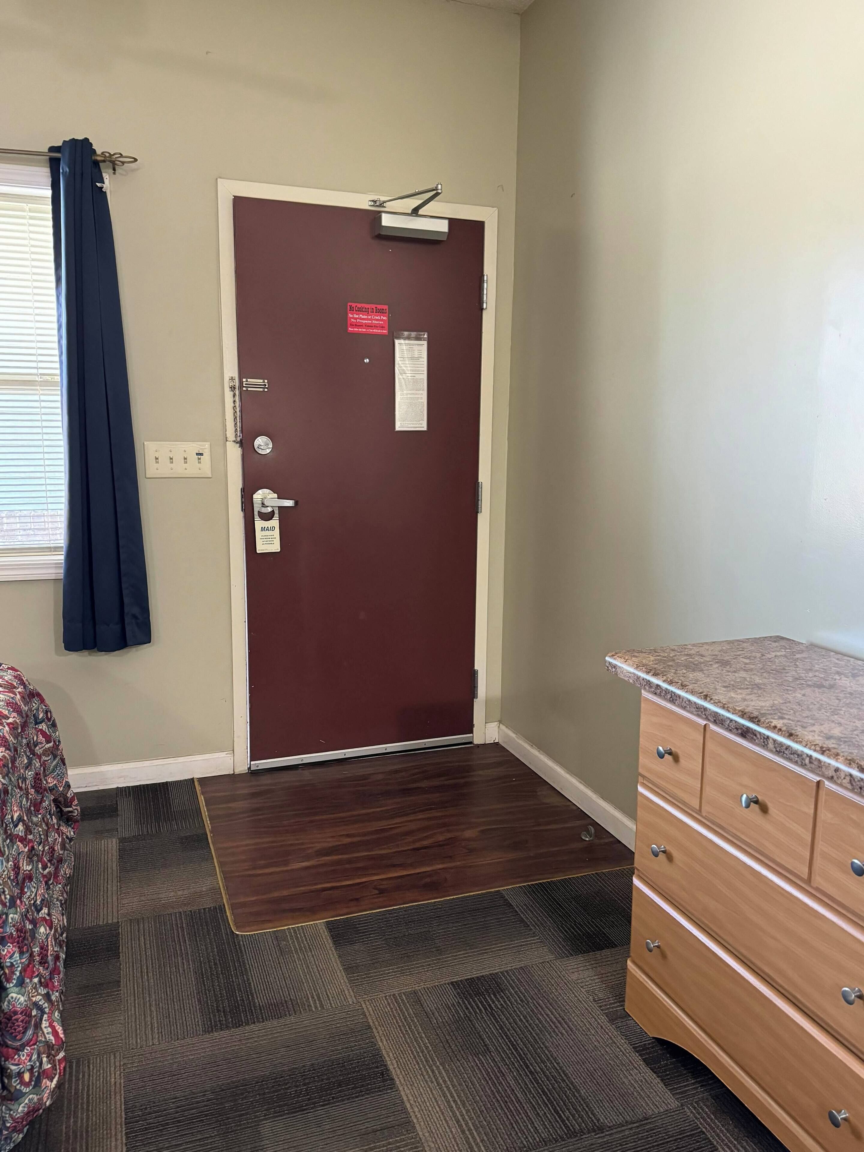 Une chambre d’hôtel avec une commode en bois, une porte ornée d’une enseigne rouge, un tapis à motifs et une fenêtre avec des rideaux.