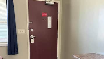 Une chambre d’hôtel avec une commode en bois, une porte ornée d’une enseigne rouge, un tapis à motifs et une fenêtre avec des rideaux.