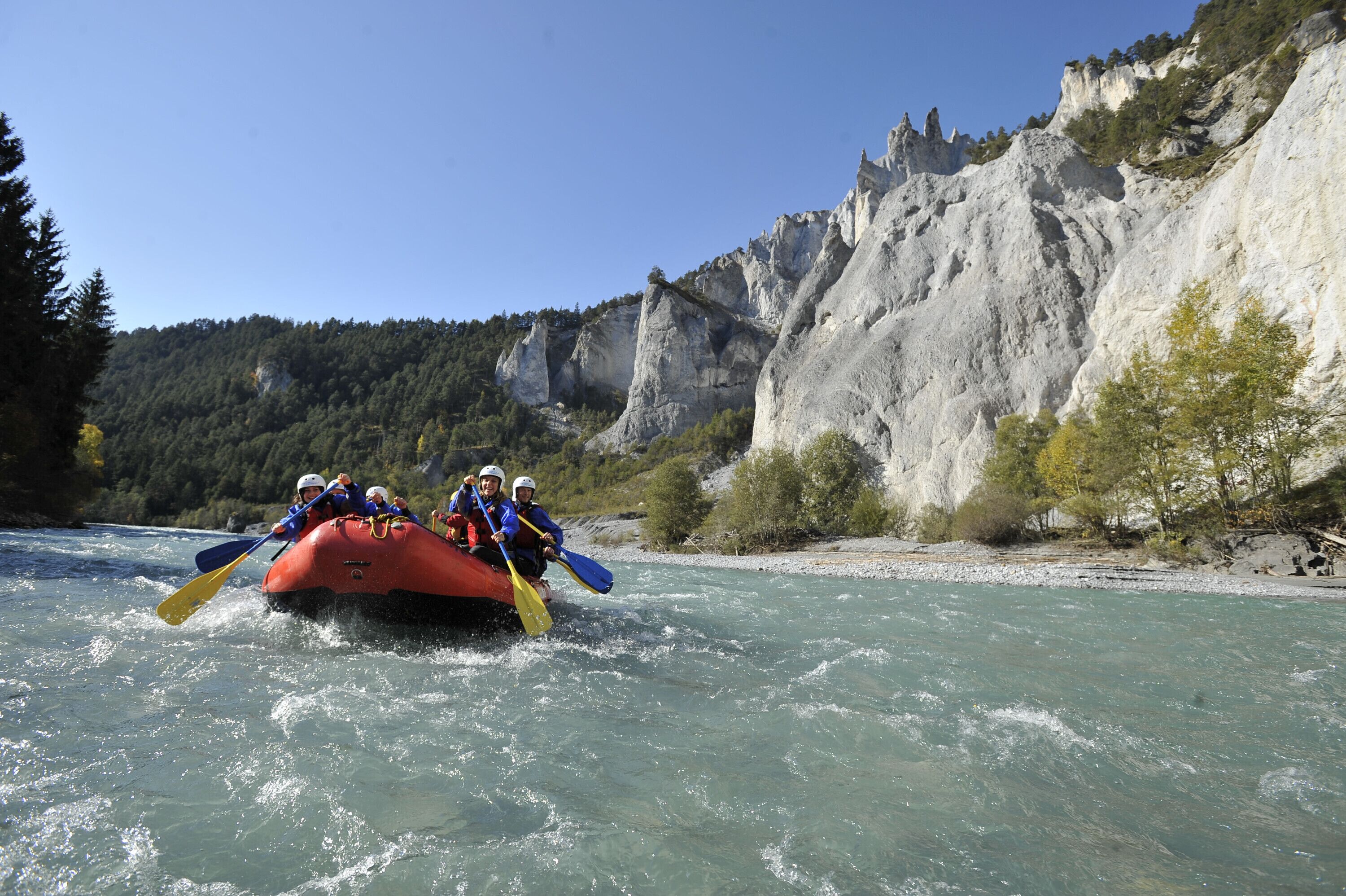 A group of people in a red raft navigating a river surrounded by rocky cliffs and trees.