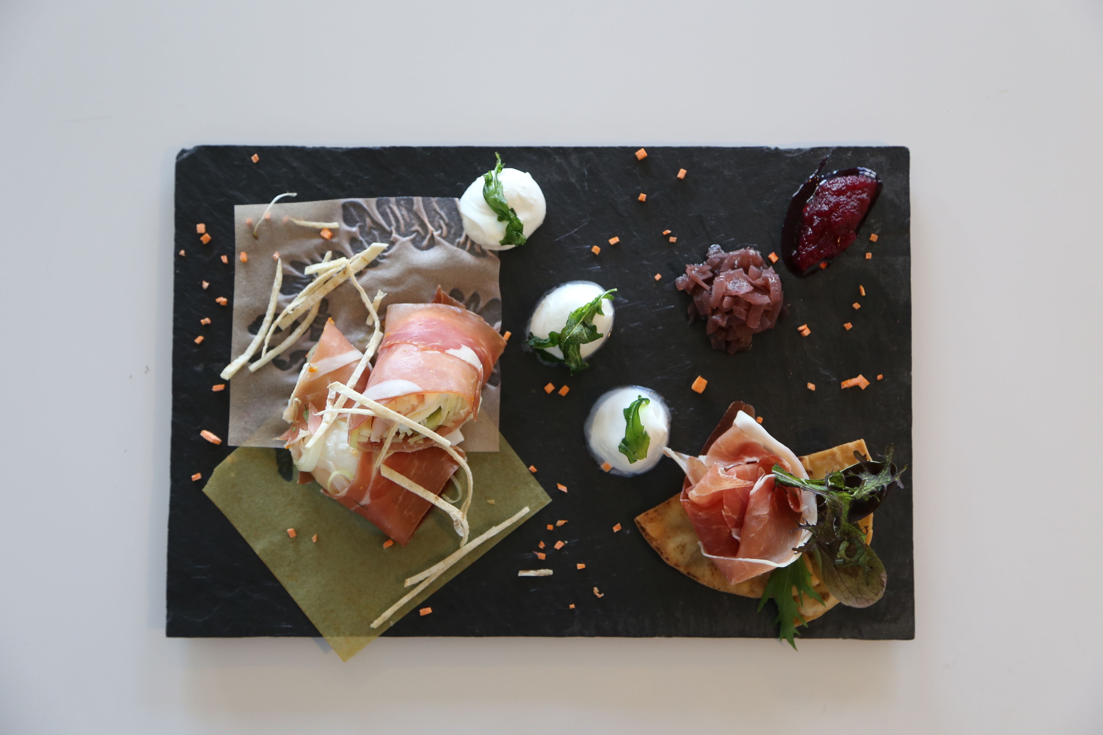 A black slate serving board with various food items including prosciutto, mozzarella, and a red fruit garnish.
