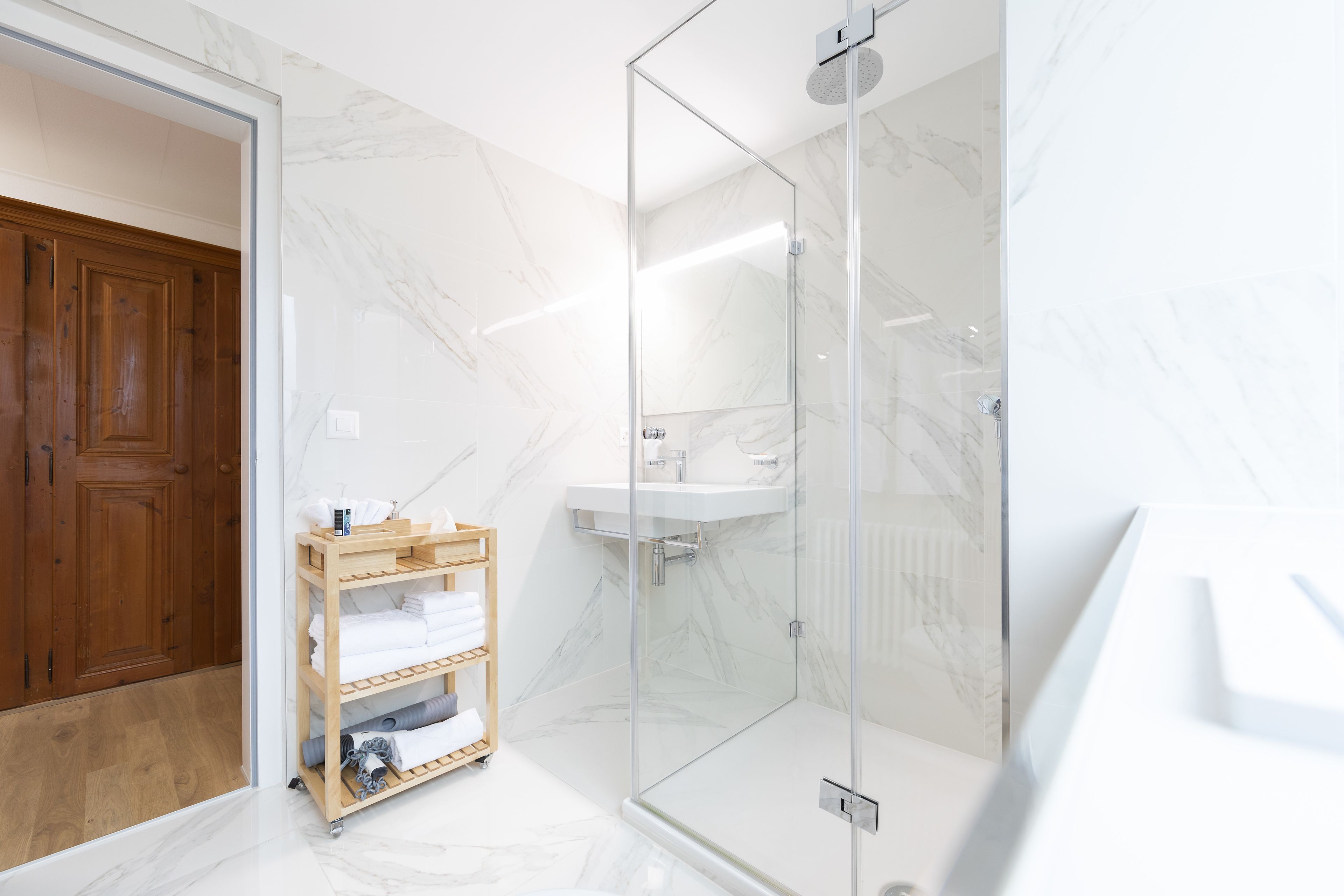 A modern bathroom with a glass-enclosed shower, a wooden shelf with towels, and a marble wall.