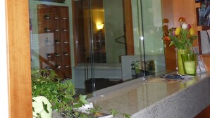 Una zona de recepción con un separador de vidrio, un mostrador de madera, un jarrón con flores y una planta en maceta.