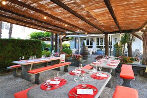Un espace de restauration en plein air avec des tables et des bancs en pierre, un toit de chaume et des plantes en pot.