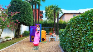 Un parque infantil con un tobogán rosa y columpios, rodeado de vegetación y un edificio al fondo.